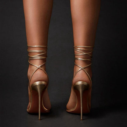 Gold ankle strap stiletto sandals with thin high heels and wrap-around ankle straps on dark background